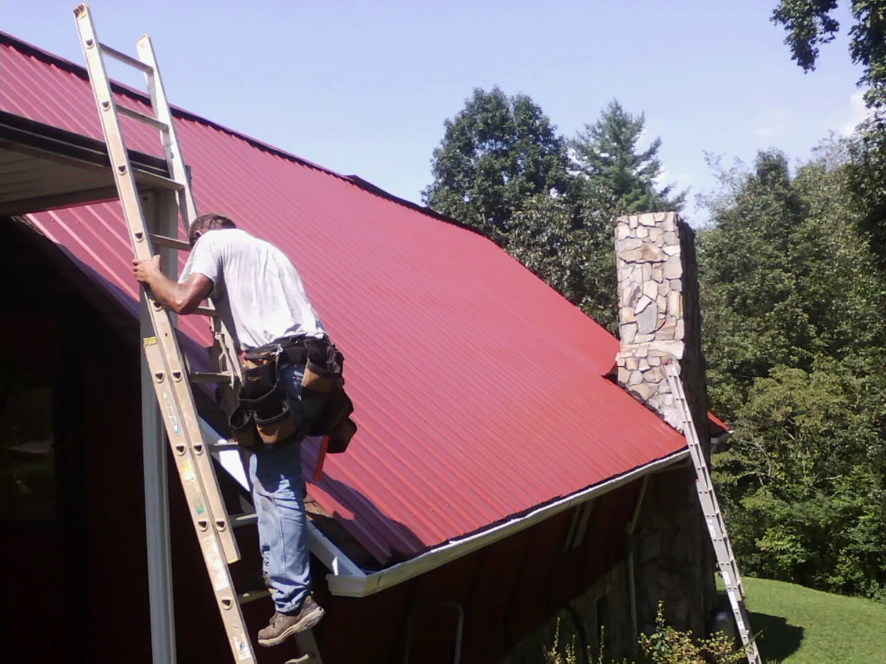 Roofer on a ladder beside a red metal roof and stone chimney — Soffit & Fascia Repair by Roofing Brothers in Commodore
